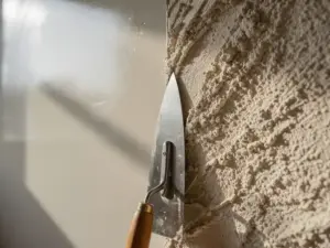 Metselspaan tegen een muur met gladde gepolijste pleister en ruwe textuurpleister naast elkaar, warme crème- en beige tinten.