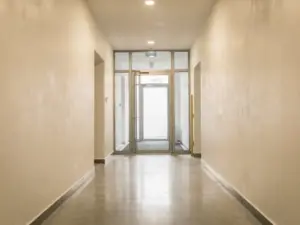 Vers gepleisterde entreehal van een appartementengebouw met gladde ivoren muren, gepolijste betonvloer en glazen voordeur.