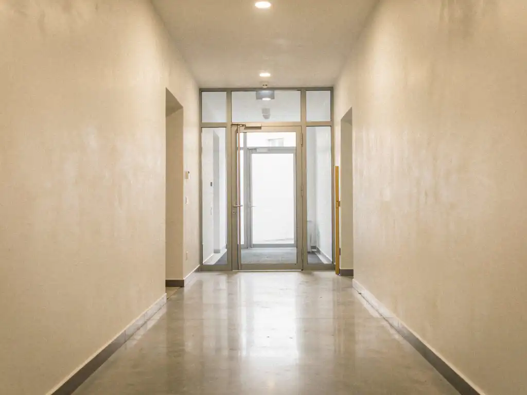 Vers gepleisterde entreehal van een appartementengebouw met gladde ivoren muren, gepolijste betonvloer en glazen voordeur.