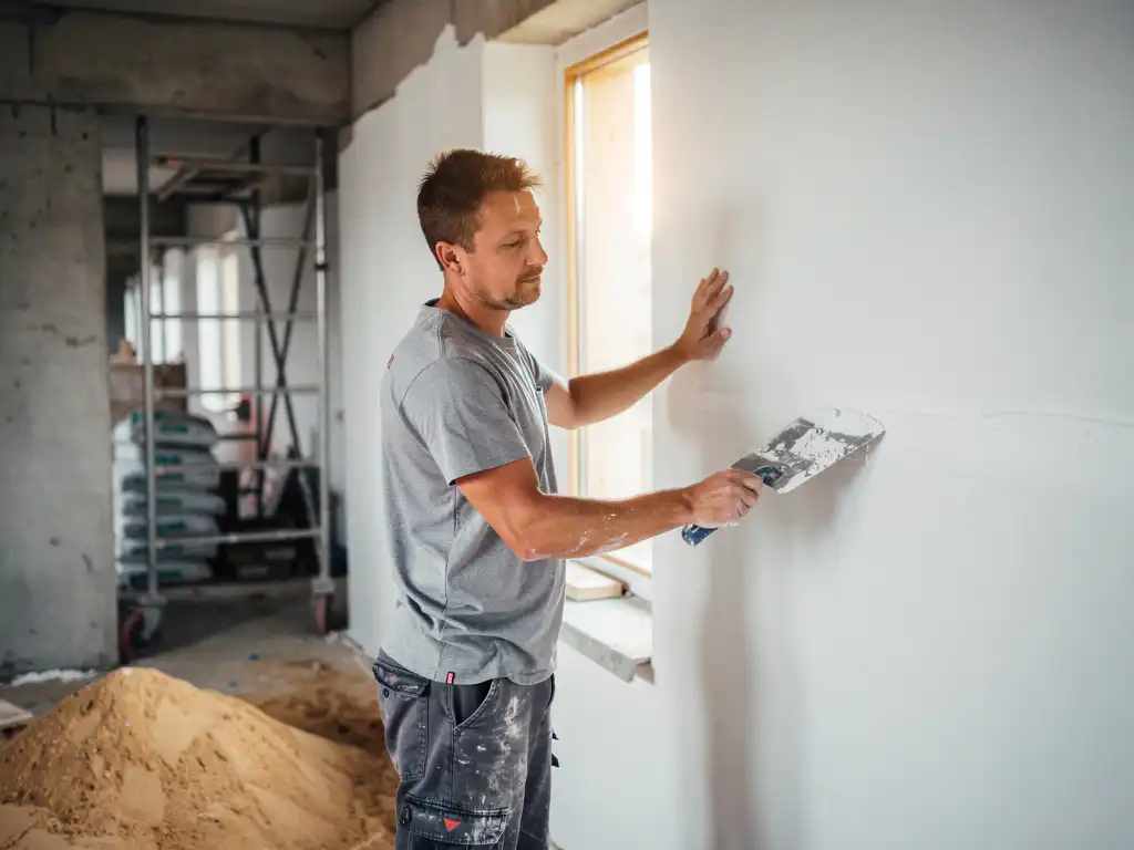 Vakman die witte stucco aanbrengt op een binnenmuur in een nieuwbouwwoning, met steigers en bouwmaterialen op de achtergrond.