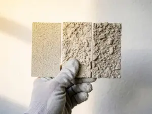Gehandschoende hand houdt drie voorbeeldplaten met fijn, middel en grof spuitpleister naast elkaar voor een witte pleistermuur.