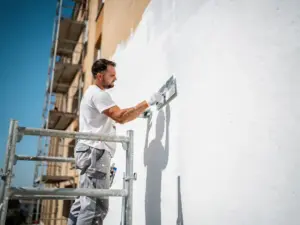Stukadoor brengt glad wit stucwerk aan op gevel van appartementengebouw, steigers zichtbaar tegen blauwe lucht.